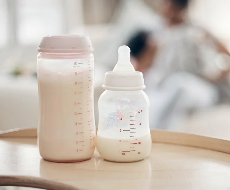 Two baby bottles filled with milk on a wooden tray, with a blurred background of a person holding a child.
