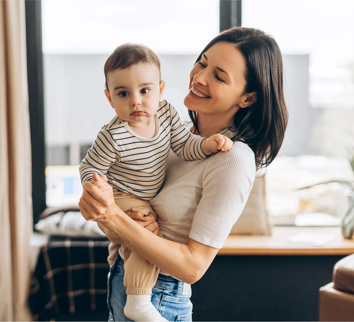 Smiling woman holding a baby wearing a striped shirt in a bright room.