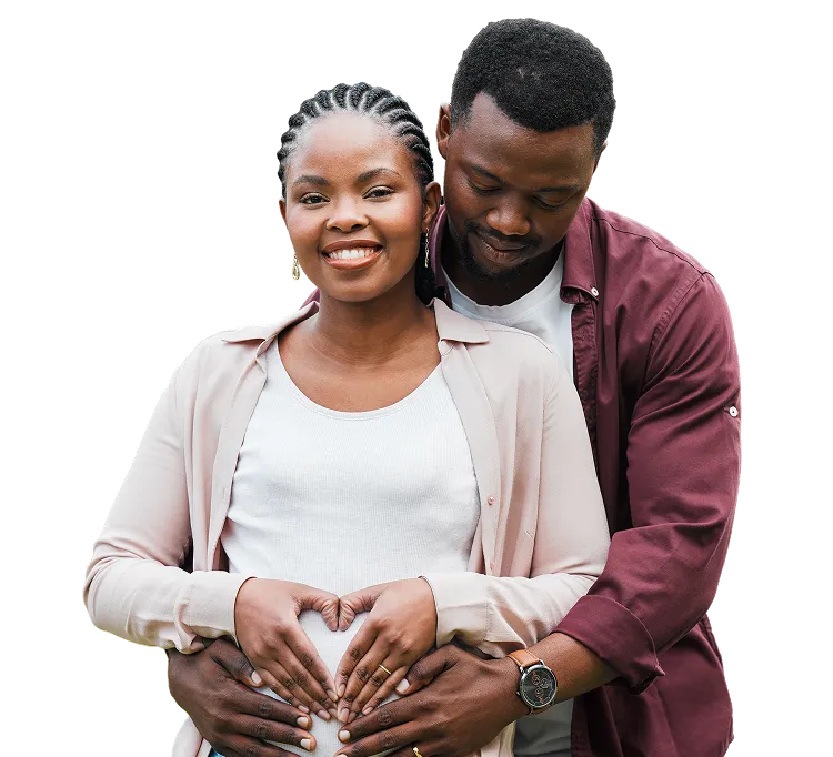 Smiling pregnant woman embraced from behind by a man, both making a heart shape with their hands over her belly.