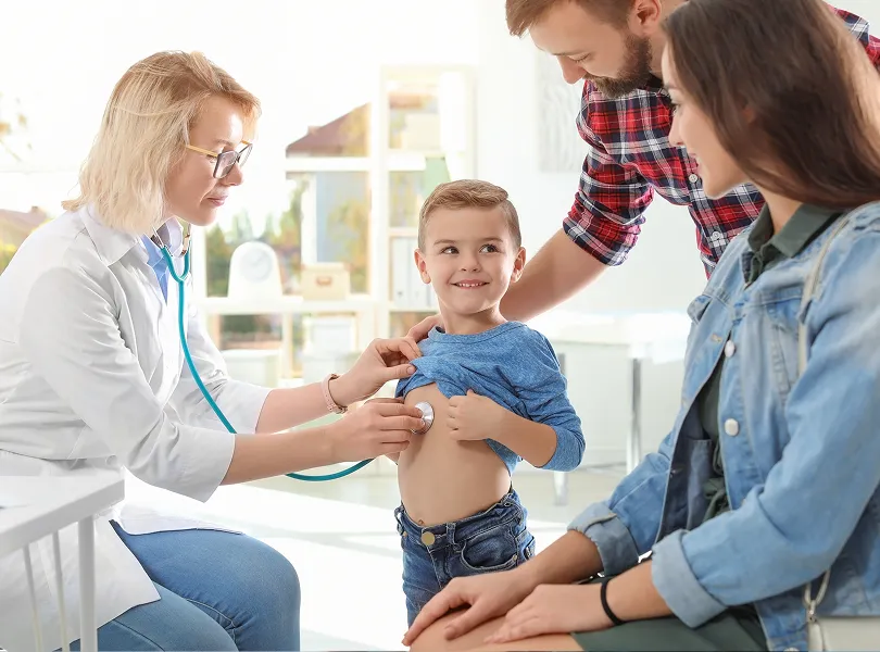 Female doctor using a stethoscope to examine a smiling young boy while his parents watch in a bright medical office.