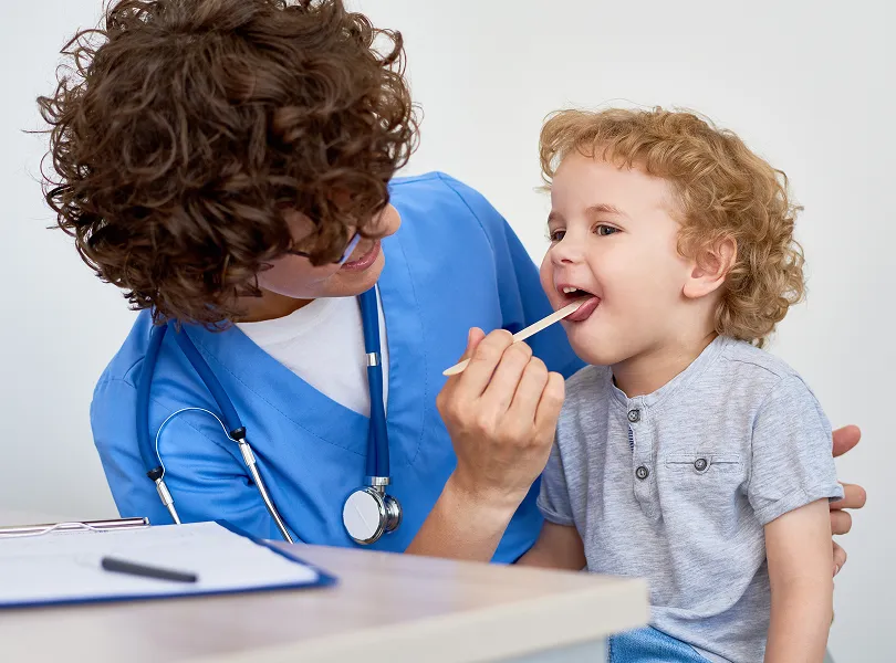 Healthcare professional in blue scrubs examining a young boy's throat with a tongue depressor.