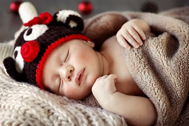 Sleeping baby wearing a crocheted ladybug hat, wrapped in a soft brown blanket on a knitted surface.