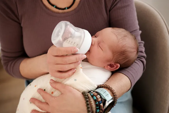 Adult holding and bottle-feeding a newborn baby wrapped in a soft blanket.