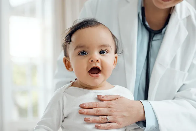 Smiling baby with open mouth sitting on a doctor’s lap during a medical checkup.