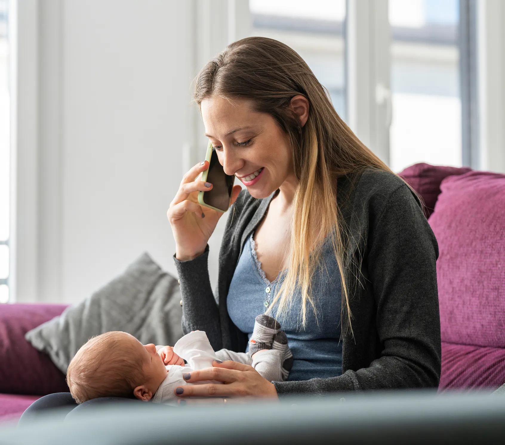 Smiling woman holding a newborn baby on her lap while talking on a smartphone in a bright living room.