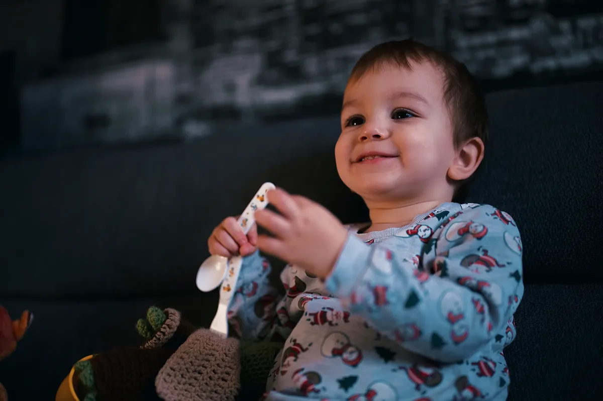 Niño sonriente en pijama de muñeco de nieve que sostiene dos cucharas de plástico mientras está sentado en un sofá oscuro.