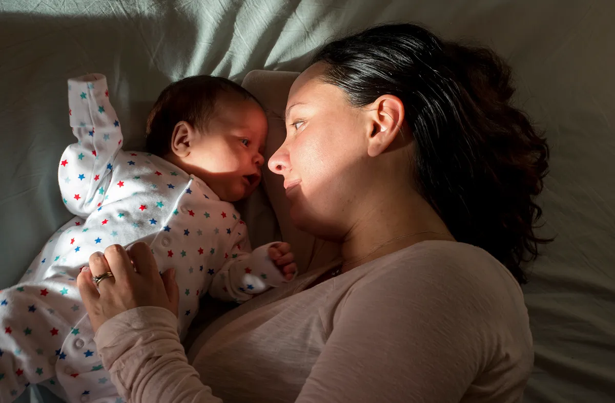 Madre acostada en la cama mirando y abrazando a su bebé recién nacido vestida con un mono con estampado de estrellas.