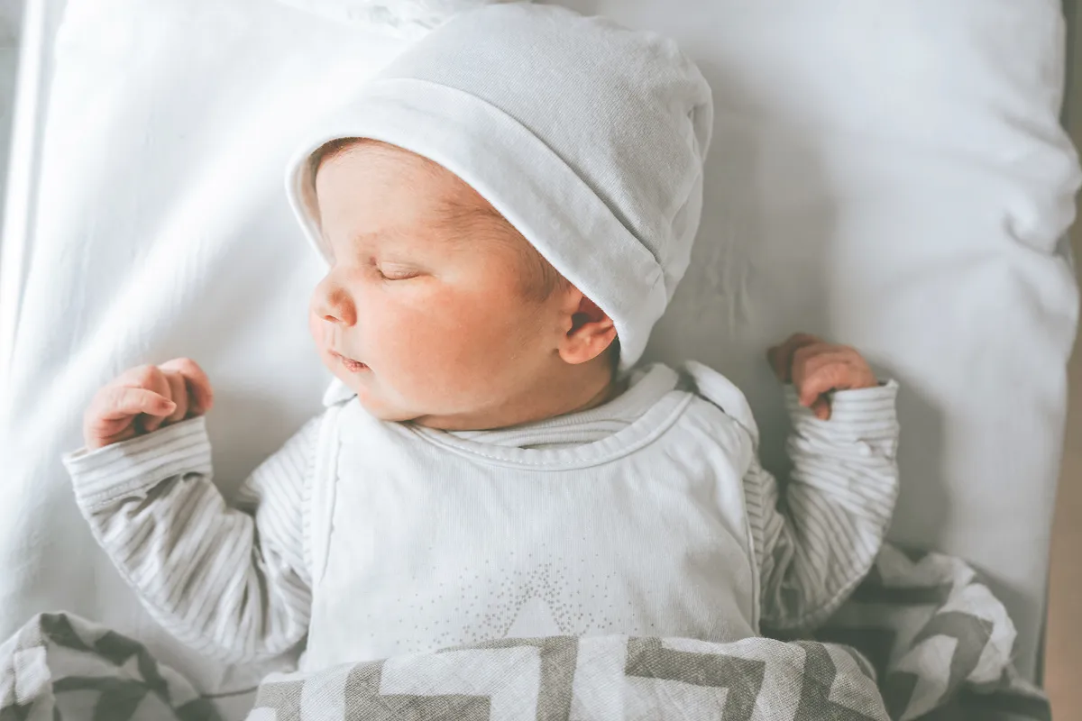 Bebé recién nacido durmiendo en una cama blanca, con un sombrero blanco y una camisa de manga larga a rayas, parcialmente cubierto con una manta estampada gris y blanca.