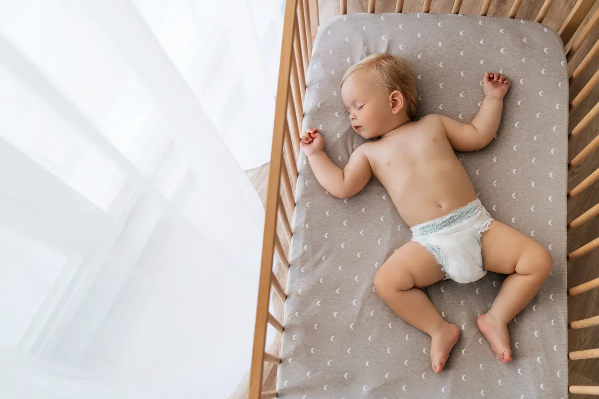 Bebé durmiendo boca arriba en una cuna de madera con sábana gris con estampado de luna.