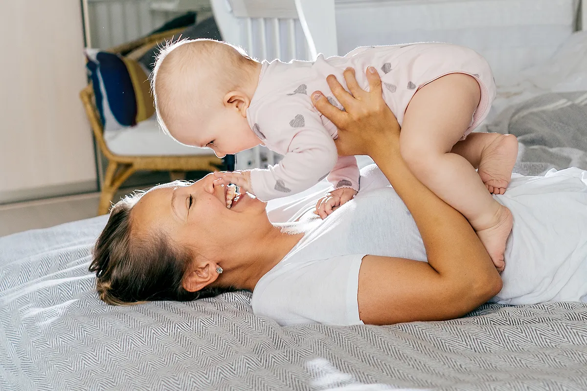 Madre acostada en la cama sosteniendo al bebé por encima de ella, sonriendo y tocándose la nariz.
