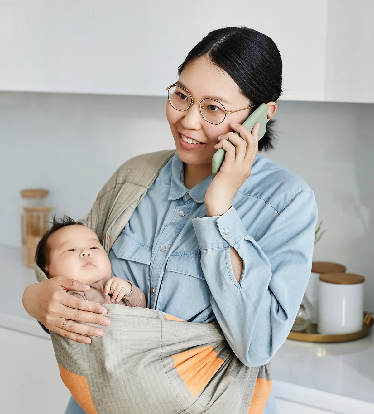 Mother wearing glasses holding a baby in a sling while talking on a phone in a kitchen.