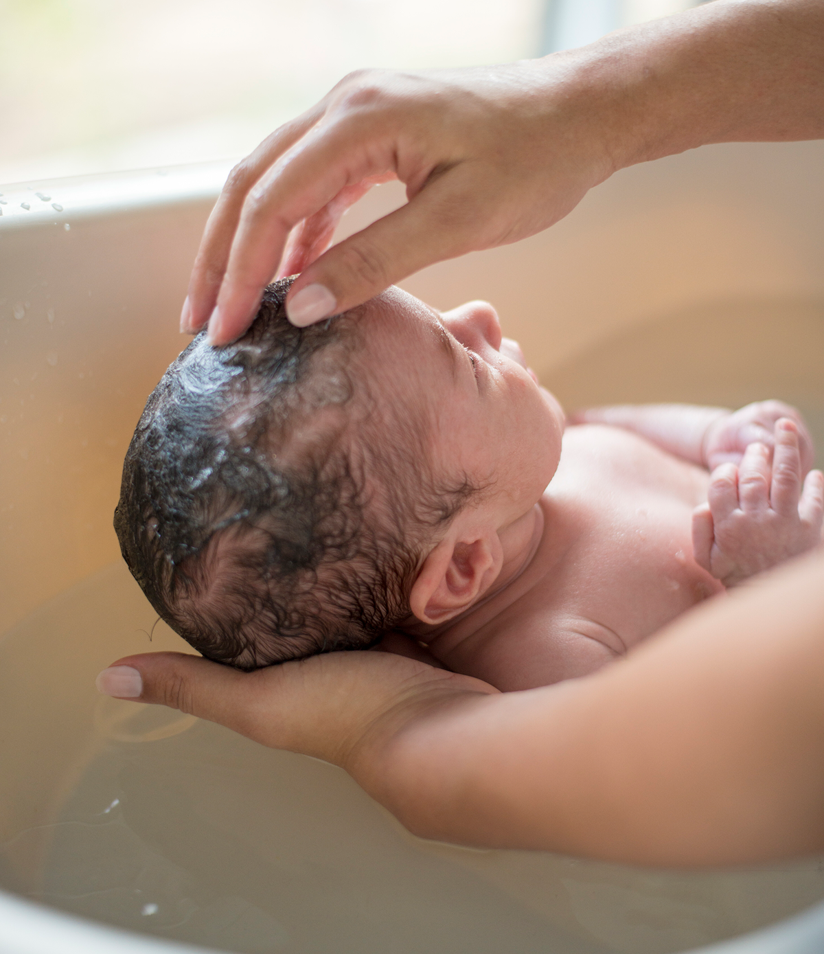 Smiling mother bathing her baby in a blue plastic tub filled with soapy water.