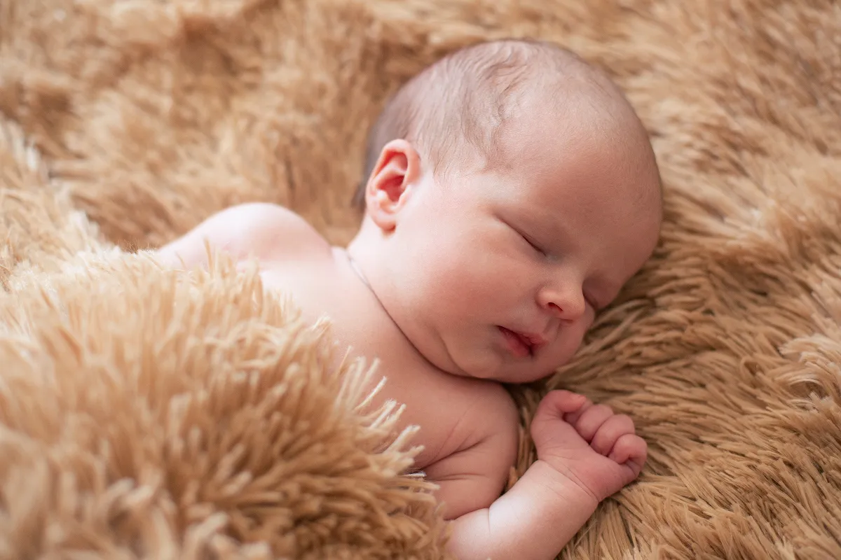 Ti bebe k ap dòmi, li gen yon onesie ble trikote sou li, kouche nan yon bèso an bwa ak dra ki gen modèl zetwal.