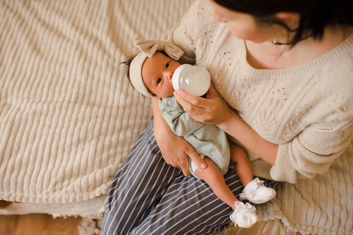 Ti bebe k ap dòmi, li gen yon onesie ble trikote sou li, kouche nan yon bèso an bwa ak dra ki gen modèl zetwal.