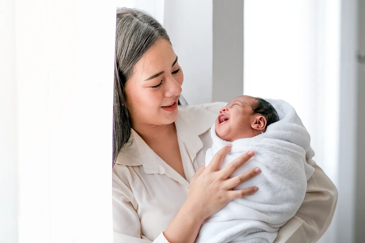 A woman sitting on a bed feeding a baby with a bottle near a large window.