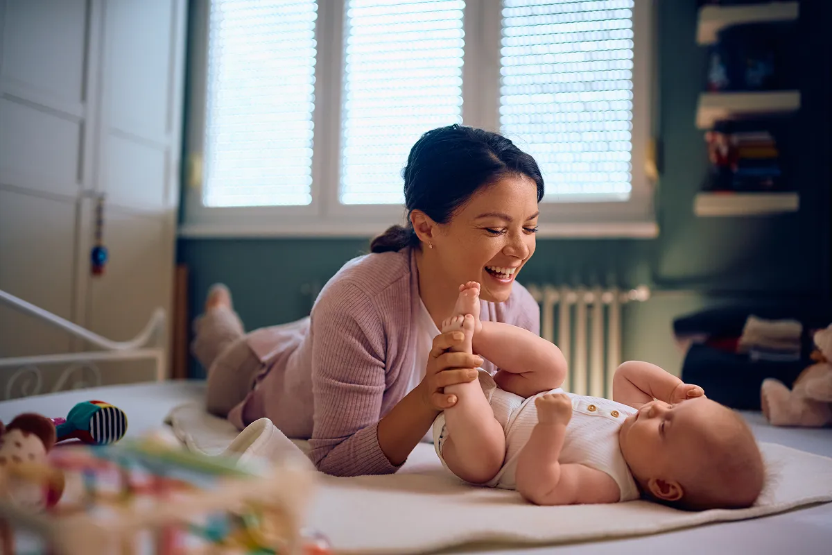 Mother lying on bed holding baby above her, both smiling and touching noses.