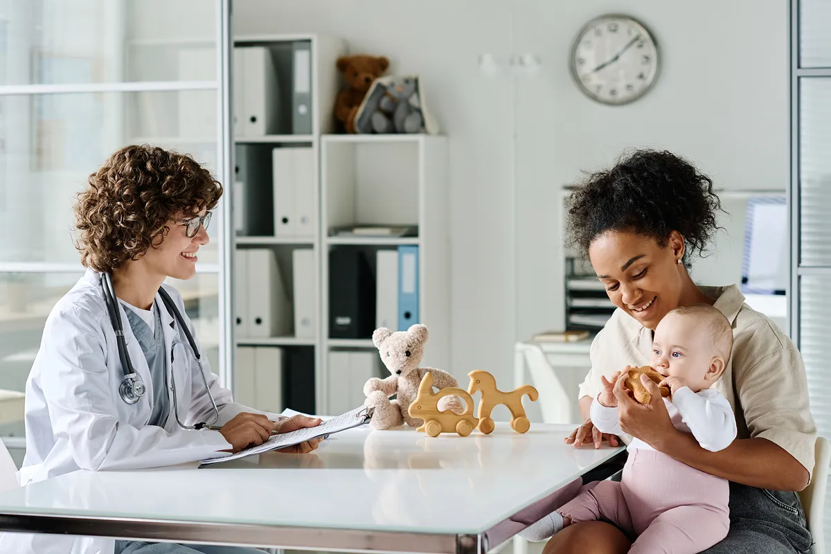 Pediatrician smiling and holding clipboard while mother and baby play with wooden toys at clinic.