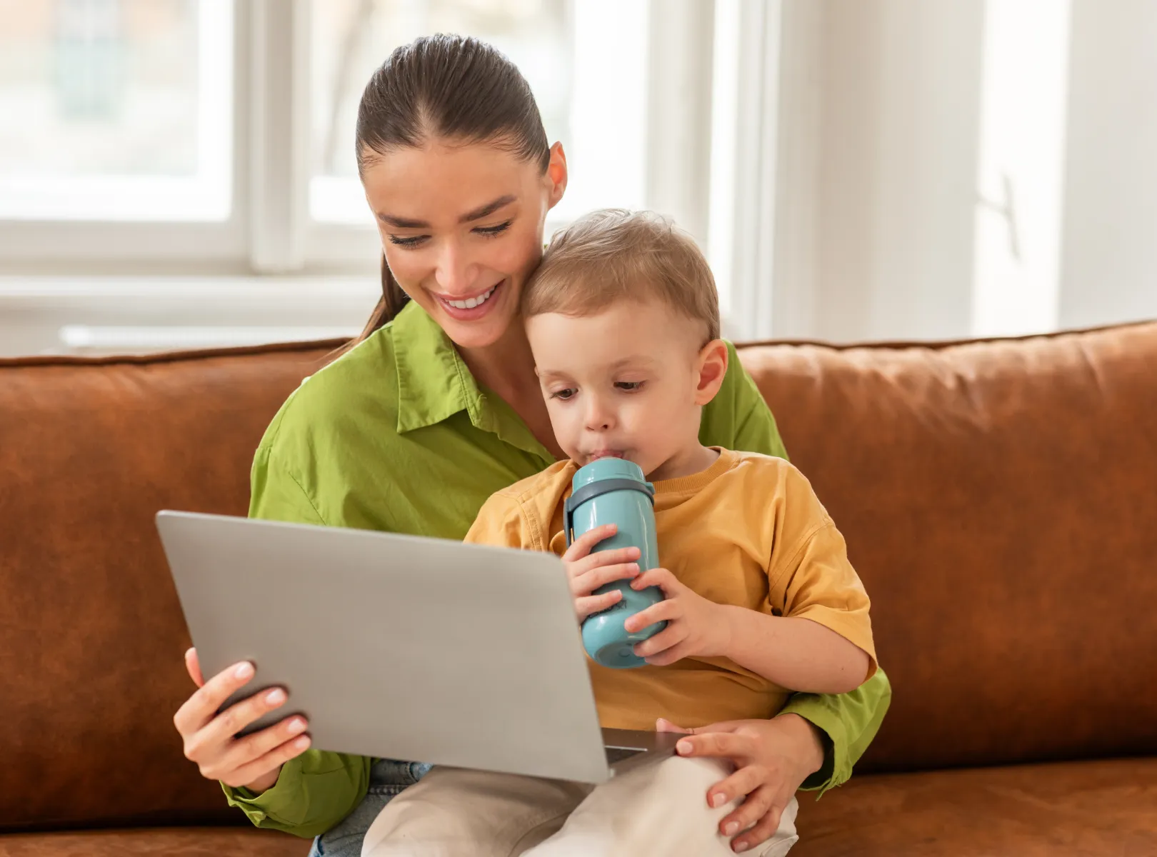 Smiling woman sitting on a couch with a toddler who is drinking from a blue sippy cup while using a laptop.