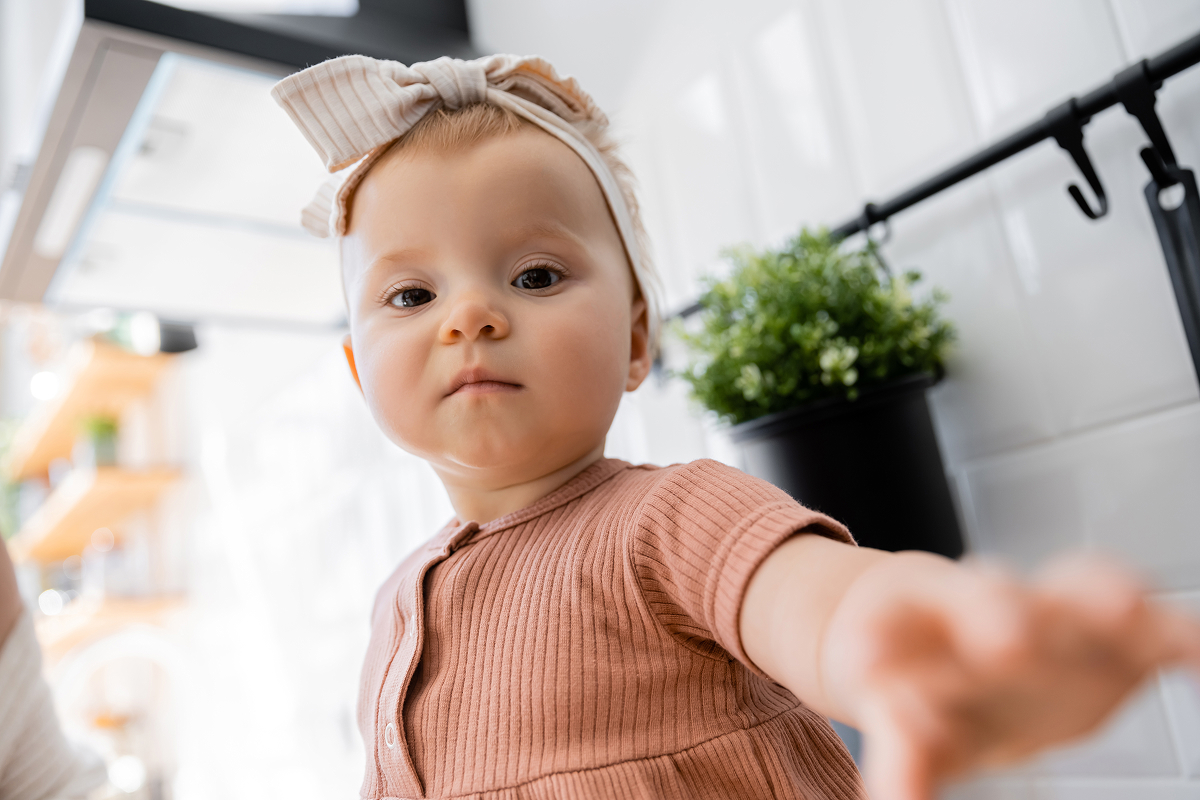 Baby girl in a bow headband reaching toward the camera in a kitchen
