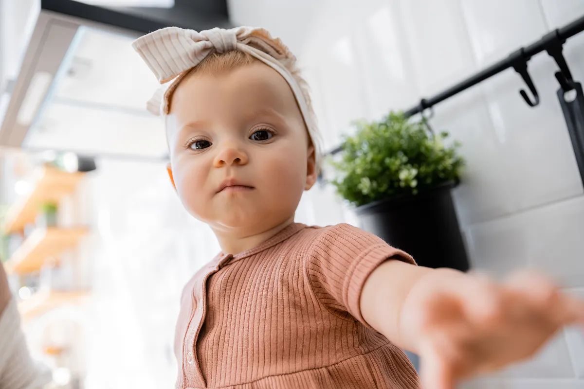 Baby girl in a bow headband reaching toward the camera in a kitchen