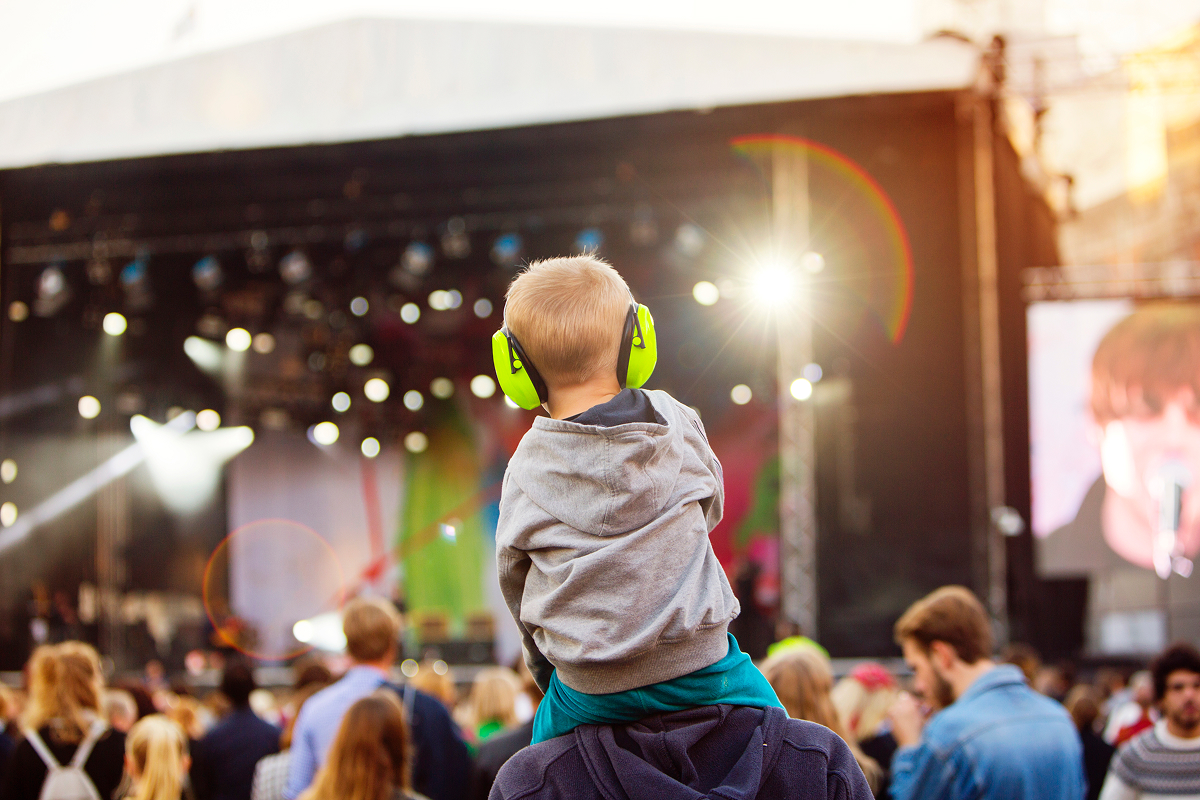 Toddler wearing bright green ear protection on an adult's shoulders facing a concert stage