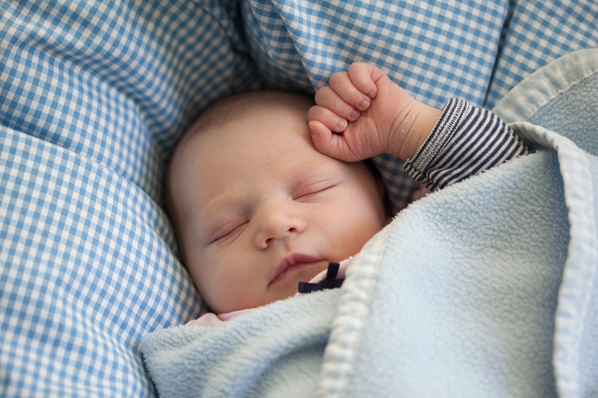 Newborn baby asleep under a light blue fleece blanket in a blue gingham bassinet