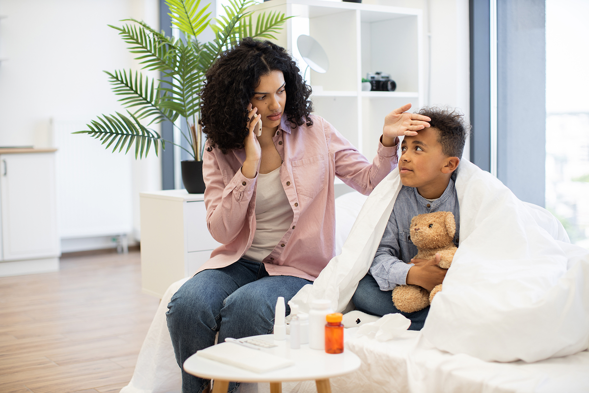 Mother on the phone checking her son's forehead as he rests on the sofa with a teddy bear and medicine on the table