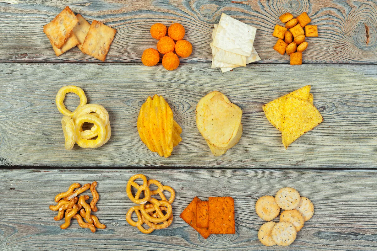 Crackers, chips, pretzels and cheese puffs laid out in rows on a weathered wooden table