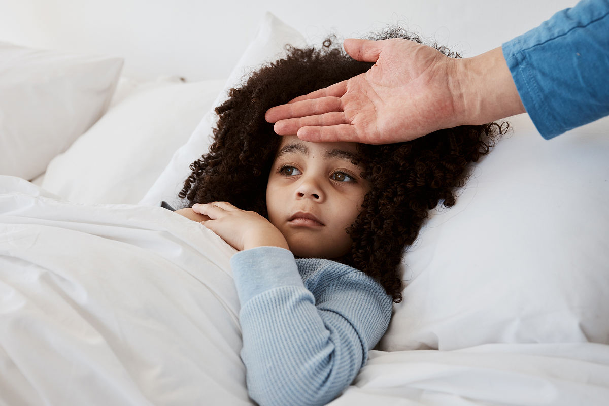Parent's hand resting on the forehead of a young girl lying in bed under white bedding