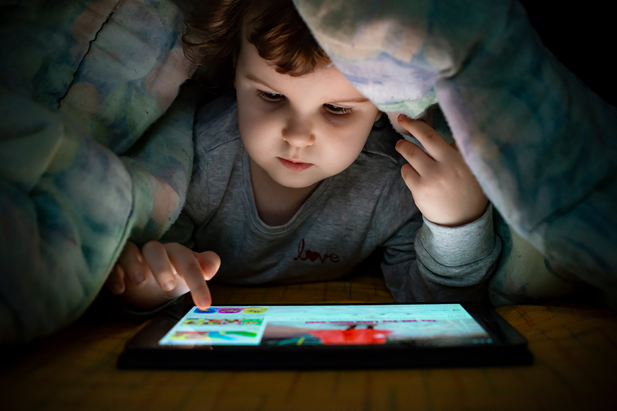 Young child hidden under a duvet at night touching the screen of a glowing tablet