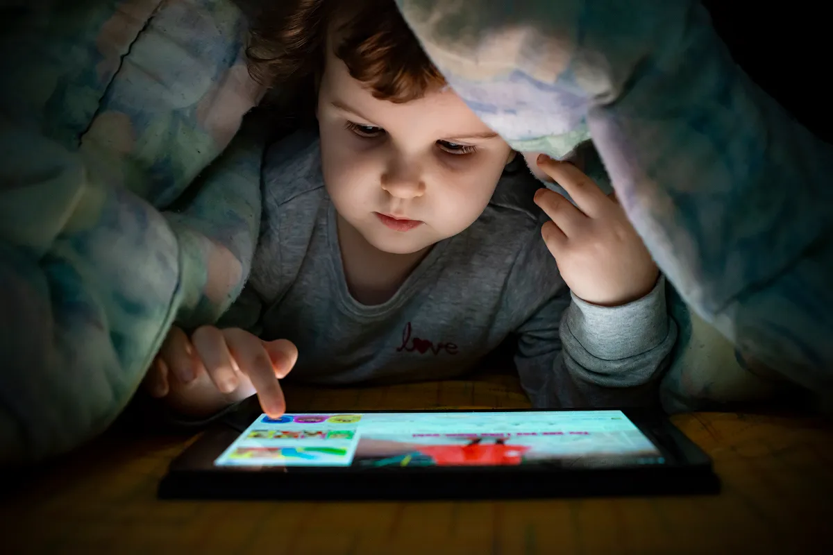 Young child hidden under a duvet at night touching the screen of a glowing tablet