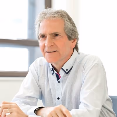 Middle-aged man with gray hair in a white shirt sitting at a table indoors, smiling slightly.