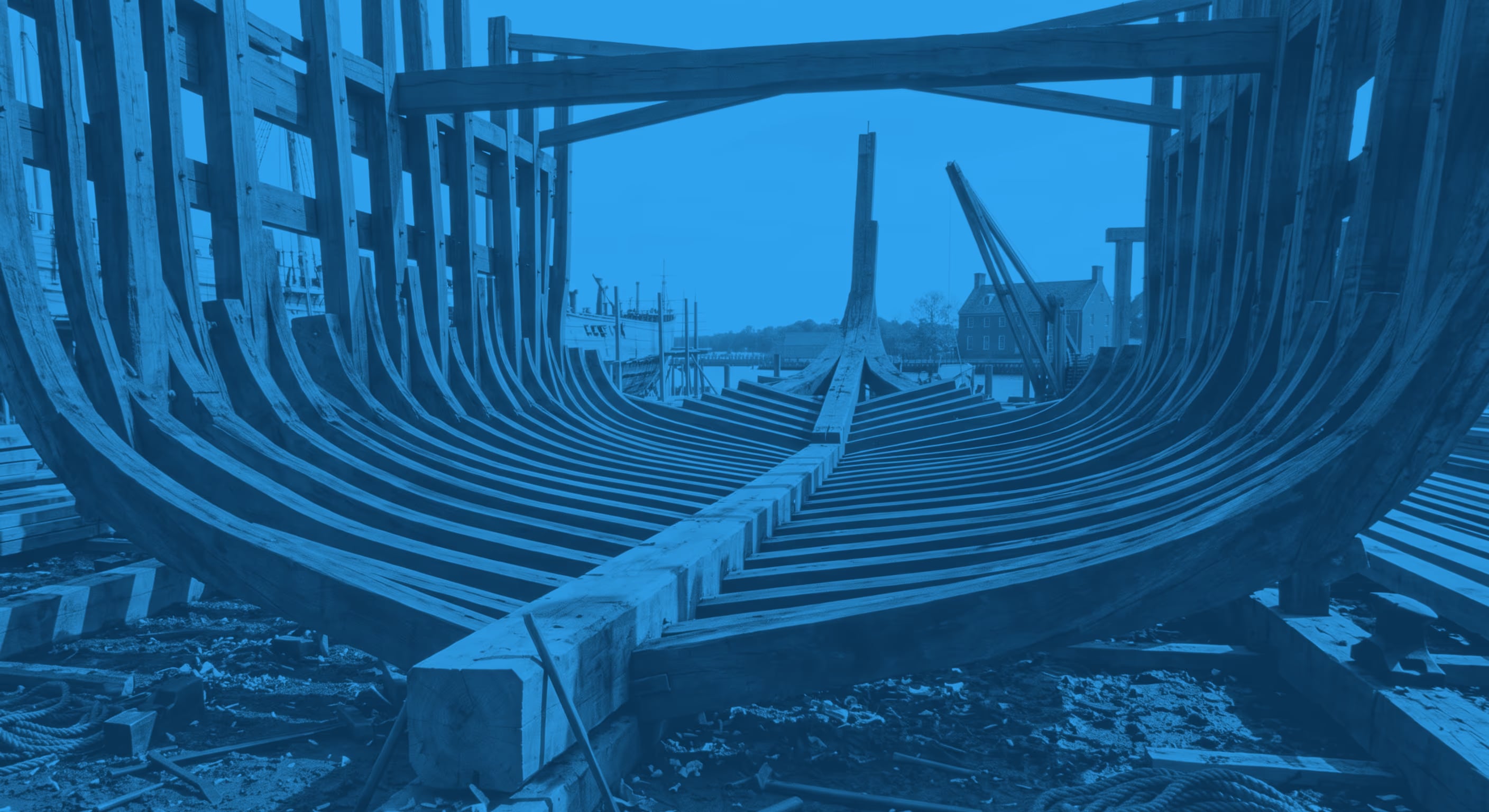The oak frames of a ship's hull standing in a shipyard, keel laid and shipwright's tools on the ground