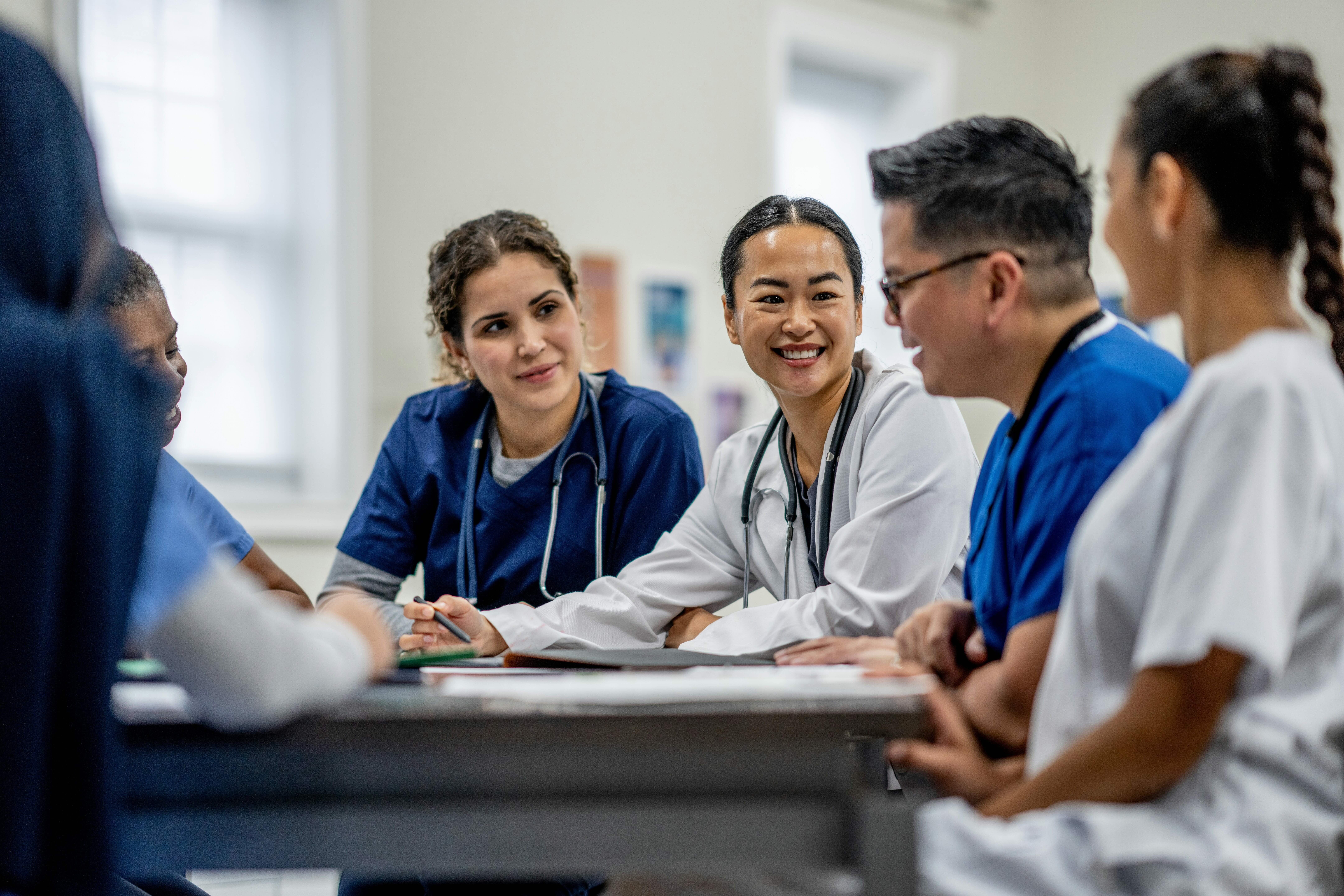 A diverse group of medical professionals in scrubs and lab coats sitting around a table, engaged in discussion and smiling.