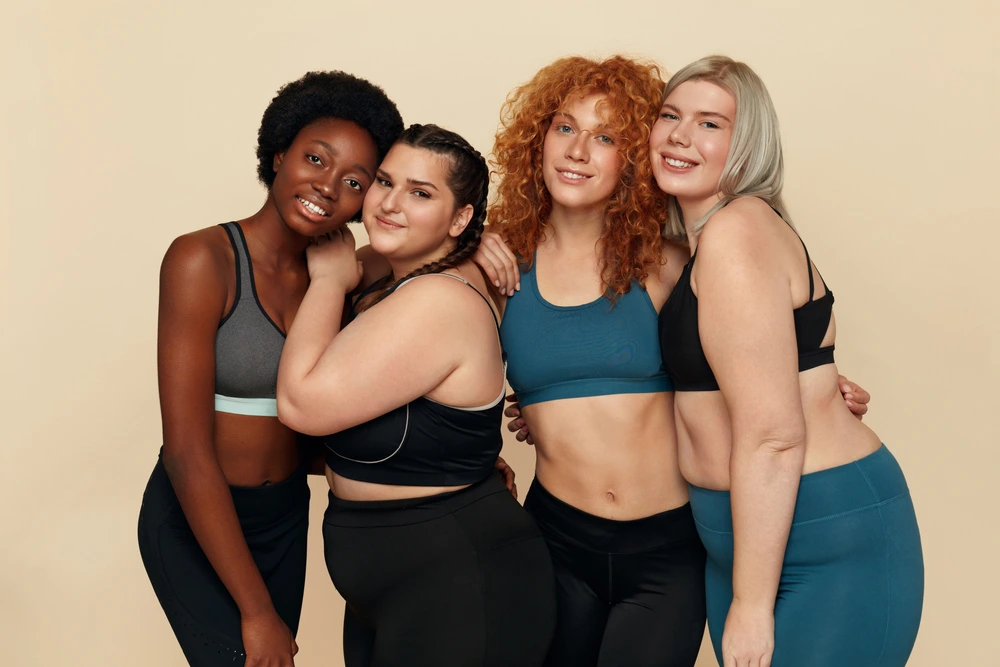 Vier Frauen in Sportkleidung zeigen unterschiedliche Körperformen und strahlen Selbstbewusstsein aus.
