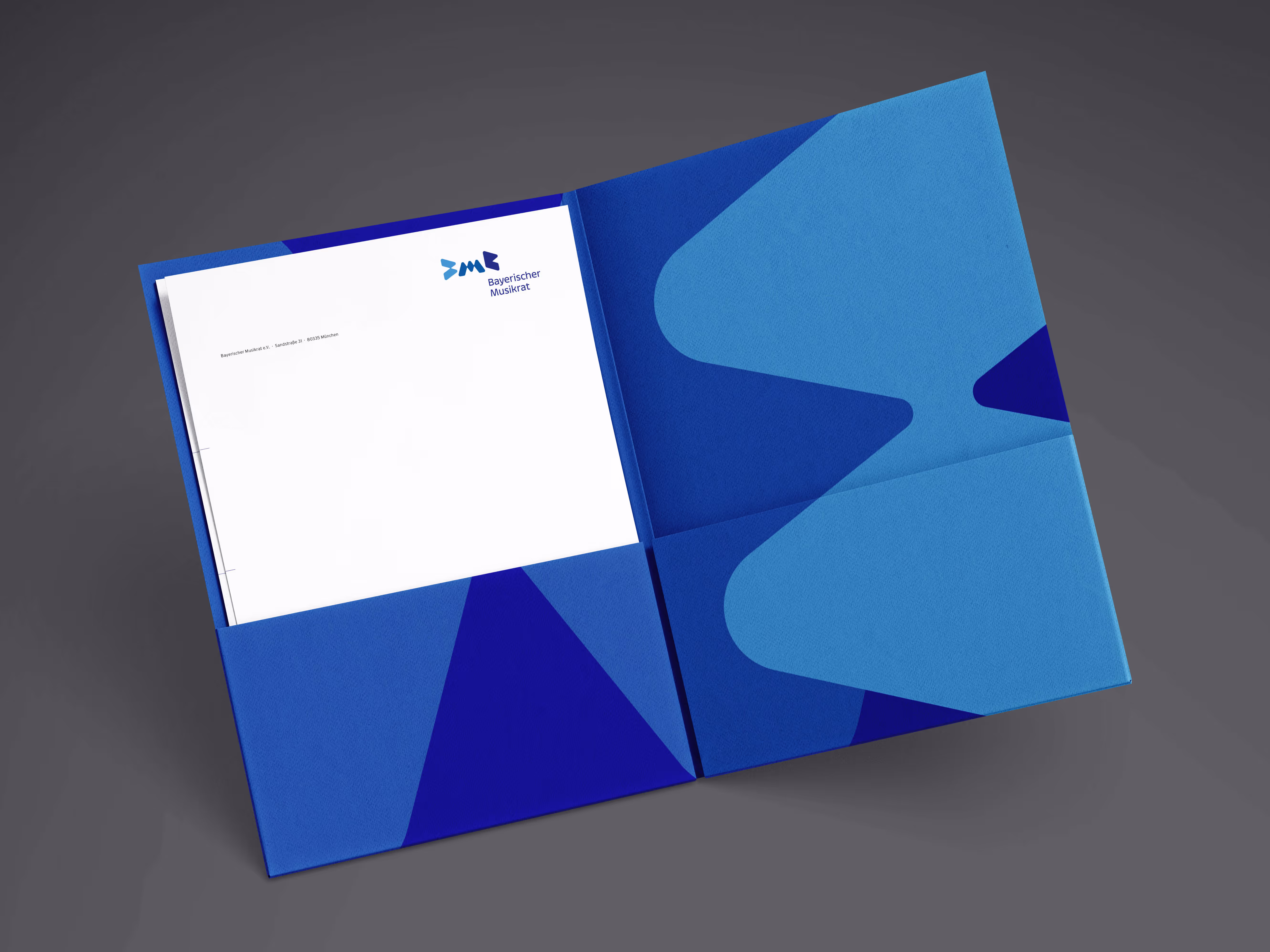 Auf einer dunklen Oberfläche liegt eine offene blaue Präsentationsmappe mit modernem geometrischem Design in verschiedenen Blautönen. In der Mappe befindet sich ein weißes Dokument mit dem Logo „Bayerischer Musikrat“ in der oberen rechten Ecke, ordentlich in einer Tasche. Das Design der Mappe umfasst abstrakte Formen, die zum Branding der Organisation passen. Die Zusammensetzung ist sauber und professionell und erinnert an eine Präsentation eines Unternehmens- oder offiziellen Dokuments.