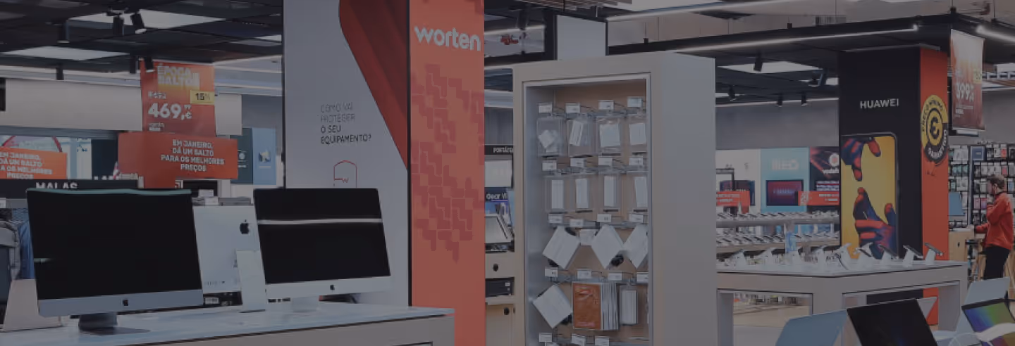 A Worten store display area featuring computers on wooden tables with promotional signage and various accessories on shelves in the background