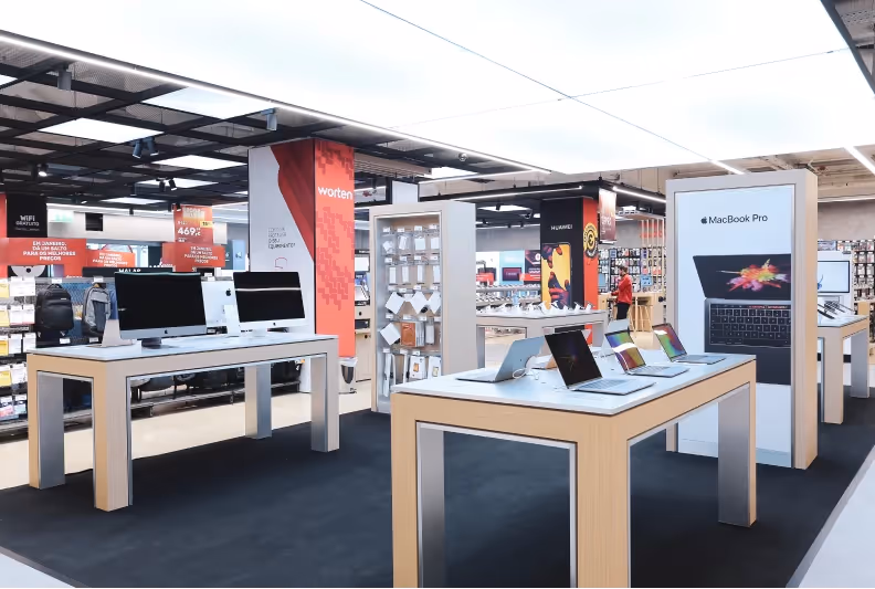 A Worten store display area featuring computers on wooden tables with promotional signage and various accessories on shelves in the background