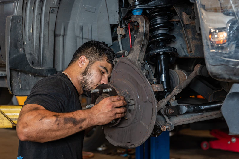 A mechanic works on a vehicle’s brake assembly, holding the wheel hub and brake rotor while inspecting the suspension and shock absorber on a car lifted in a repair shop.