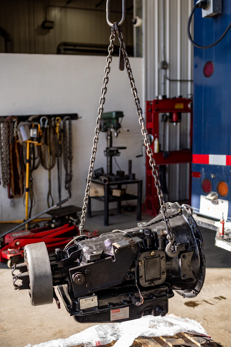 Transmission repair with heavy-duty gearbox suspended by chain hoist in shop bay during removal and service.