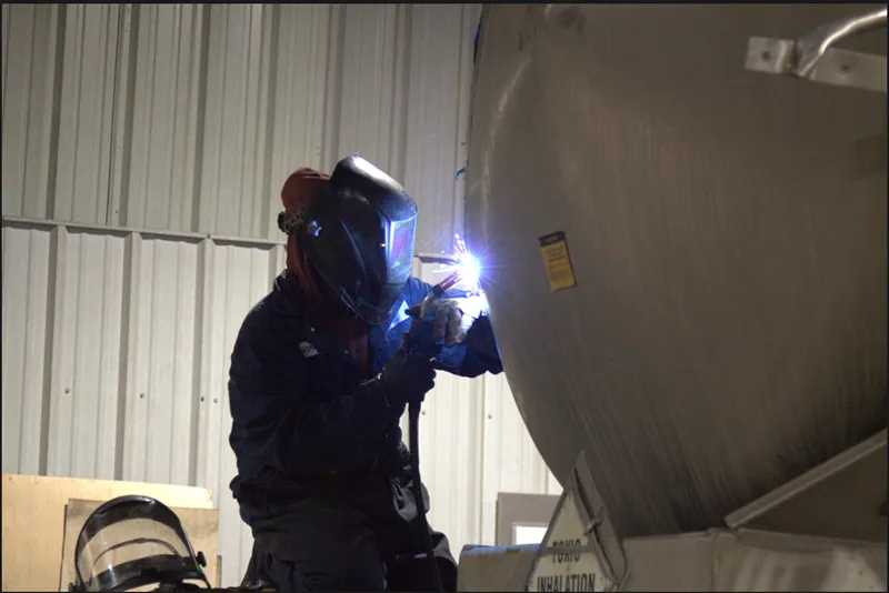 Haz-mat tank welding repair performed by a technician inside a heavy-duty shop. Yellow label text is visible on the tank.