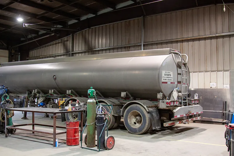 Silver tanker trailer in a heavy-duty repair shop, surrounded by welding equipment and gas cylinders.