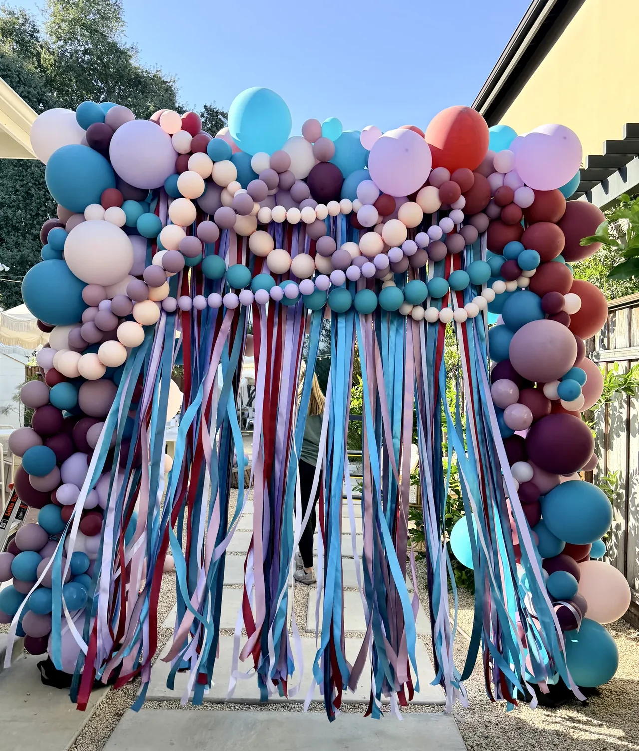 A full-size custom balloon installation in teal, lavender, blush and burgundy with flowing ribbons.