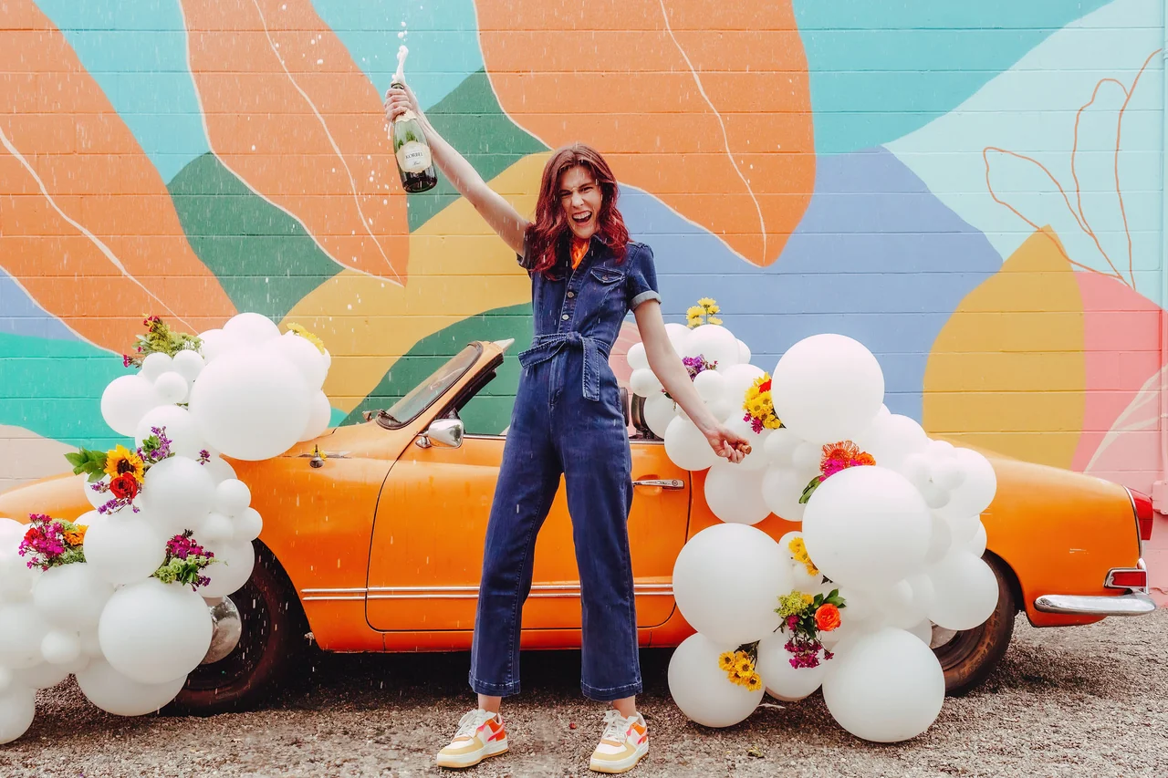 Karlie celebrating beside an orange convertible decorated with white balloons and flowers.