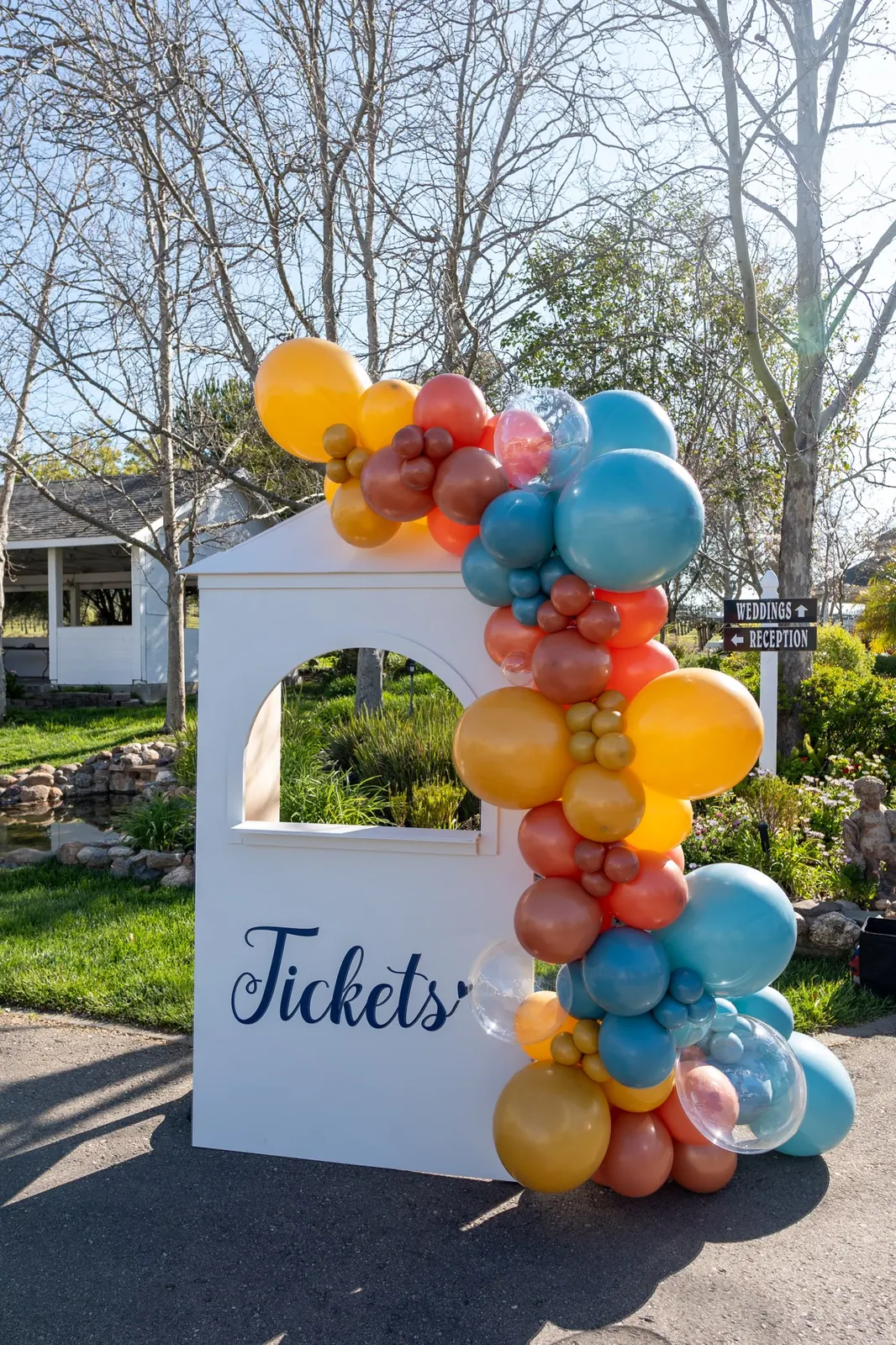 Teal, mustard, and rust balloon garland climbing a white ticket booth at an outdoor train-themed birthday