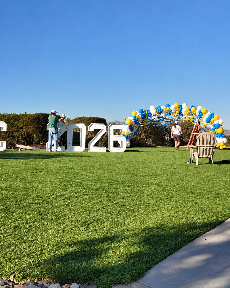 Crew placing white HOCO 2026 marquee letters beside the balloon arch on a sunny lawn