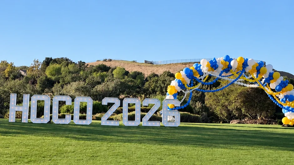 HOCO 2026 marquee letters and a blue, yellow and white balloon arch side by side on a lawn, ready for group photos