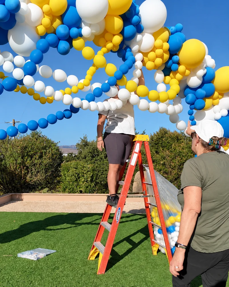 Crew member on a ladder tying white, yellow and blue link balloons inside a homecoming balloon arch