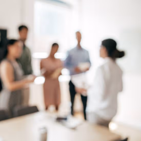 A group of five people stand in a brightly lit office, having a discussion. The image is slightly out of focus, emphasising the collaborative environment. One person faces the group, holding a piece of paper. 
