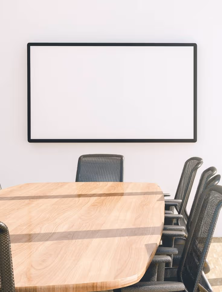 A conference room with a wooden table surrounded by black mesh chairs. A large blank whiteboard is mounted on the white wall in the background. Sunlight casts shadows across the table and floor.